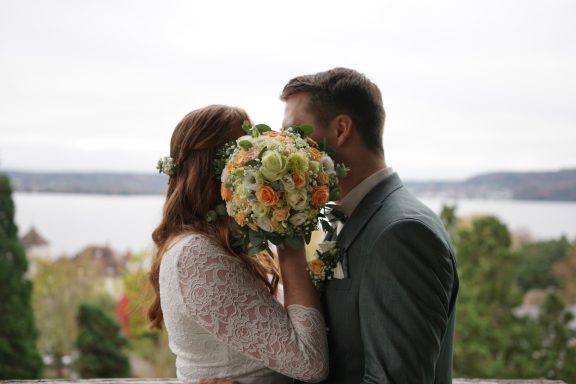 Ein junges Brautpaar mit einen Blumenstrauß vor den Gesichtern. Im Hintergrund ist der Bodensee zu sehen.